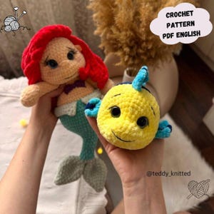 May include: Crocheted mermaid doll with red hair, a purple bow, and a teal tail, held next to a yellow crocheted fish with blue fins. The image includes the text "CROCHET PATTERN PDF ENGLISH".
