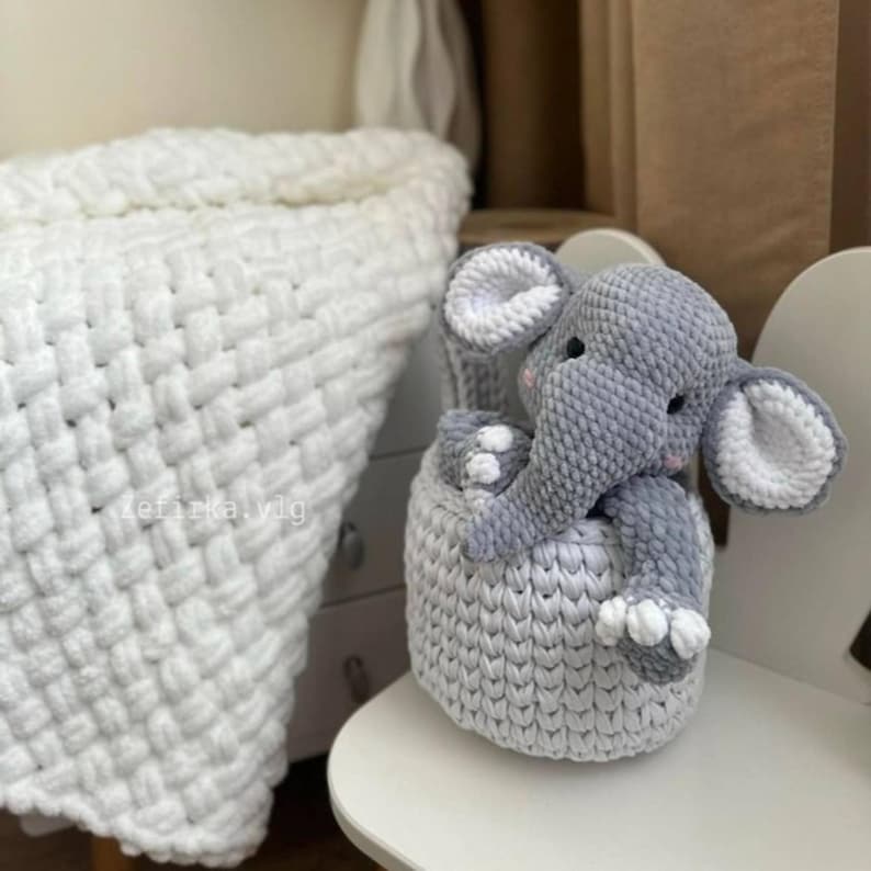 May include: A gray and white crocheted elephant toy sitting in a matching basket. The elephant has large ears with white inner ears and small pink accents. The basket and elephant are on a white chair, with a white woven blanket in the background.