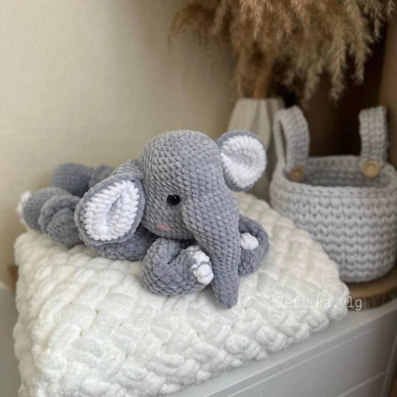 May include: A handmade gray crochet elephant toy with white accents, lying on a white textured blanket. The elephant has large ears and a trunk. A gray crochet basket with wooden buttons is in the background.