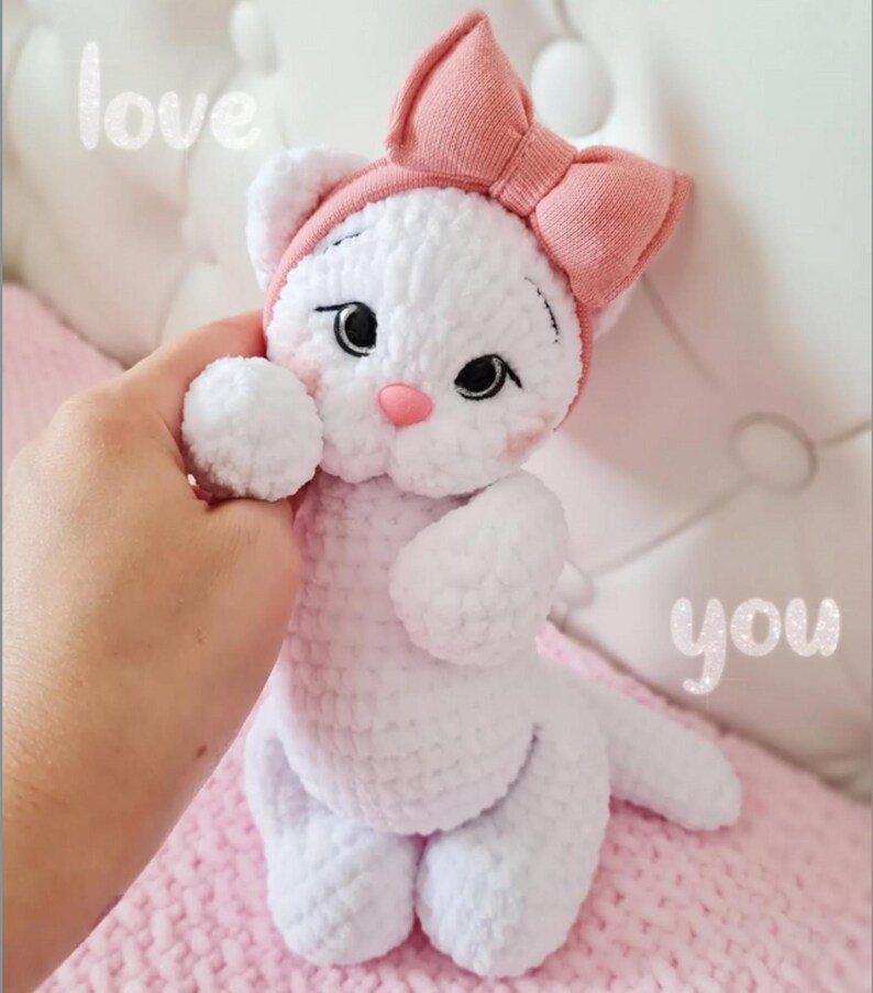Puede incluir: Un gato blanco de ganchillo hecho a mano con un lazo y una nariz rosas. El gato tiene ojos negros y est&aacute; sostenido por una mano. Las palabras "love" y "you" est&aacute;n escritas en la imagen. El juguete est&aacute; hecho de hilo blanco suave.