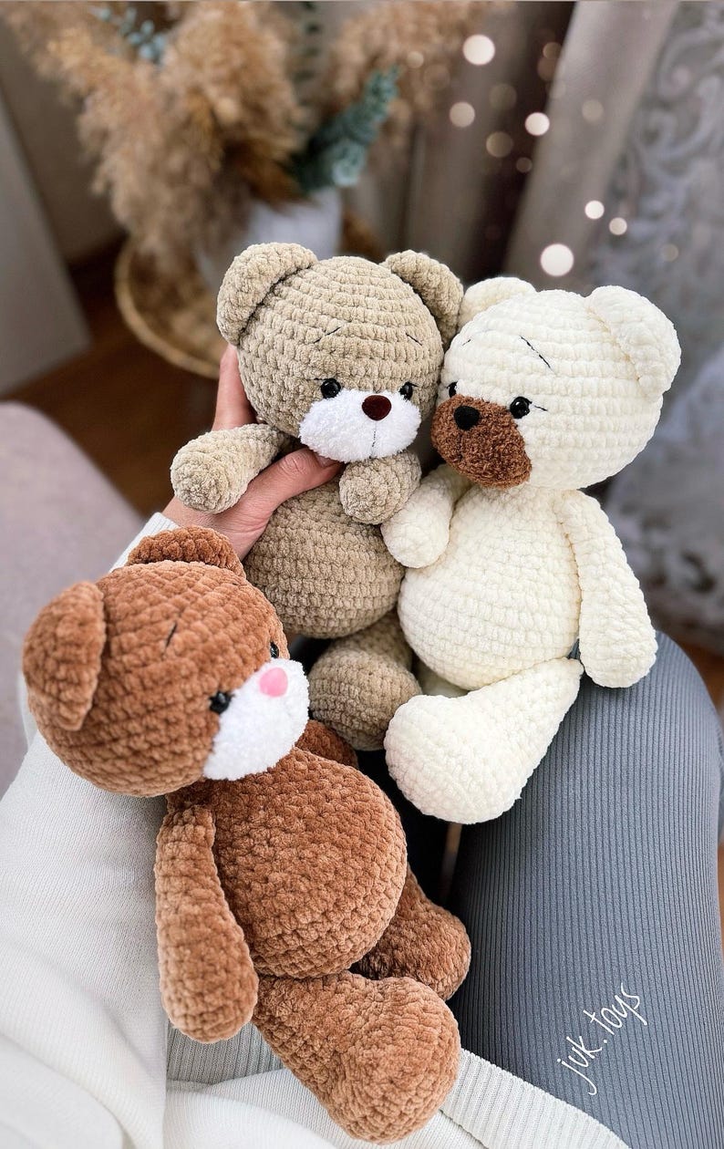 Op de afbeelding: Drie gehaakte teddyberen in verschillende kleuren: bruin, beige en wit. De beren hebben zwarte ogen en witte neuzen. De beren zitten op een grijs oppervlak met de tekst "jyk.toys" in wit.