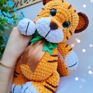 May include: A handmade orange and brown crochet tiger plush toy with white accents. The tiger has large, expressive eyes, brown stripes, and a green and brown ribbon around its neck. The plush is soft and cuddly.