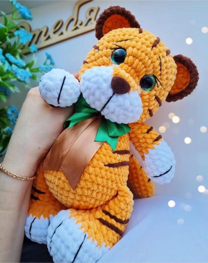 May include: A handmade orange and brown crochet tiger plush toy with white accents. The tiger has large, expressive eyes, brown stripes, and a green and brown ribbon around its neck. The plush is soft and cuddly.
