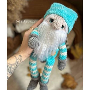 May include: A handmade crochet gnome doll with a light blue hat and striped outfit. The gnome has a long white beard, gray gloves, and gray shoes. The text "Zefirka.vlg" is visible on the image.