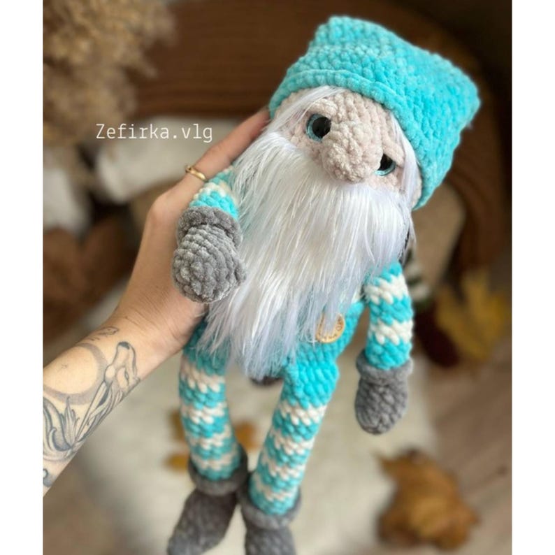 May include: A handmade crochet gnome doll with a light blue hat and striped outfit. The gnome has a long white beard, gray gloves, and gray shoes. The text "Zefirka.vlg" is visible on the image.