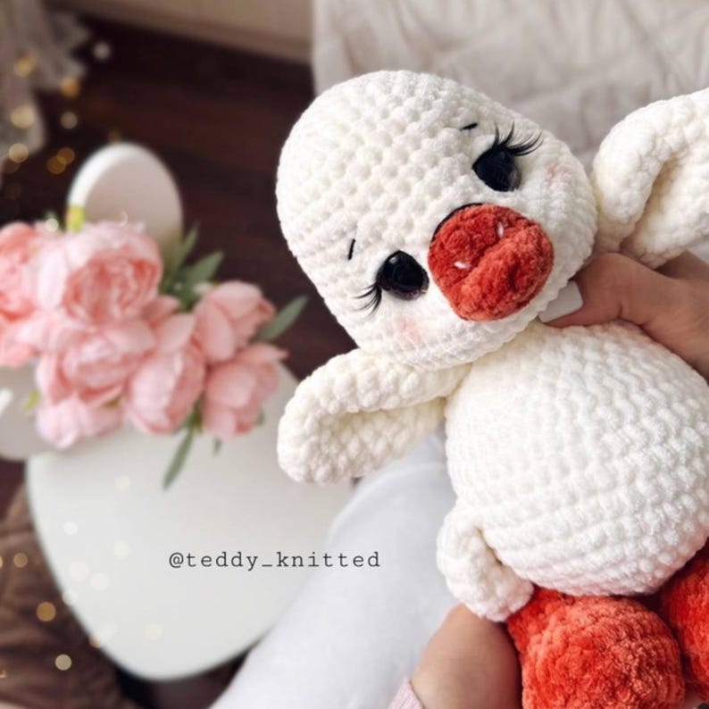 May include: A handmade white crochet duck with a red beak and feet, and black eyes with long eyelashes. The duck is being held by a person. The text "@teddy_knitted" is visible.