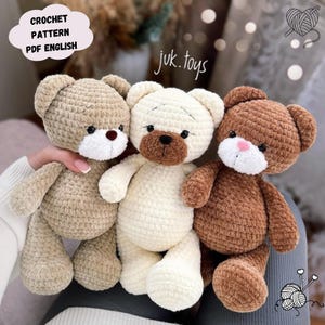 Op de afbeelding: Drie gehaakte teddyberen in verschillende kleuren: bruin, beige en wit. De beren hebben zwarte ogen, roze neuzen en witte vacht rond hun mond. De beren zitten op een grijs oppervlak. De tekst "juk.toys" is zichtbaar op de achtergrond.