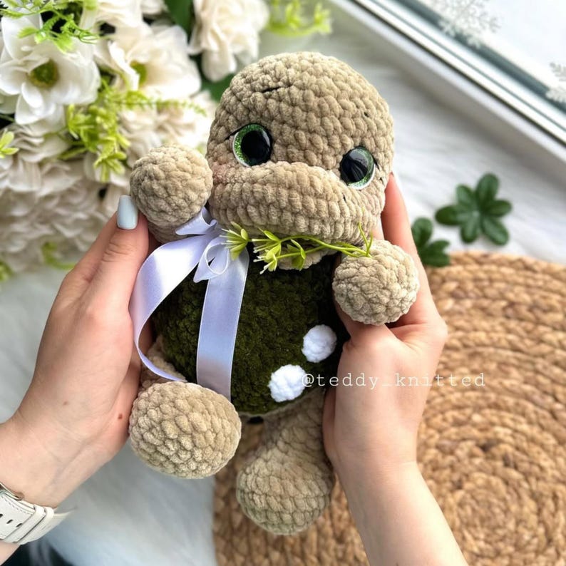 May include: A crocheted plush turtle toy with green eyes, a white ribbon, and green leaves in its mouth. The turtle is sitting in the hands of a person.