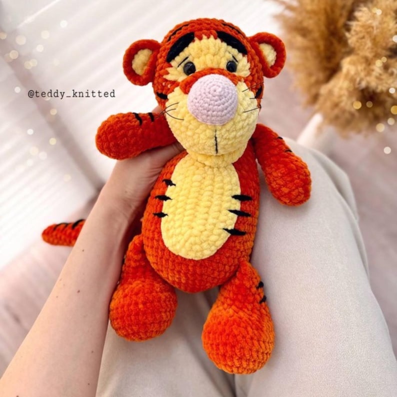 May include: A crocheted orange and yellow tiger plush toy with black stripes. The toy has a pink nose and black eyes. The toy is being held by a hand.