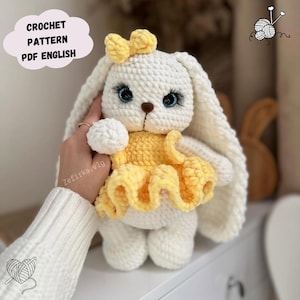 Puede incluir: Conejo de crochet blanco con un vestido amarillo y un lazo blanco. El conejo tiene ojos azules y una nariz de crochet. El conejo está sentado en una superficie blanca.