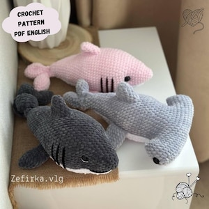 Könnte beinhalten: Drei gehäkelte Hai-Plüschtiere in Rosa, Grau und Dunkelgrau. Die Haie haben schwarze Augen und weiße Bäuche. Der Text "CROCHET PATTERN PDF ENGLISH" befindet sich in einer wolkenförmigen Blase. Die Website "Zefirka.vlg" ist ebenfalls sichtbar.
