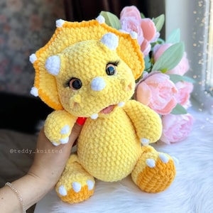 May include: A crocheted yellow triceratops stuffed animal with white horns and eyelashes. The toy is sitting on a white fluffy surface.
