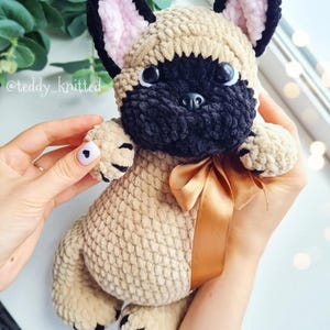 May include: A crocheted French bulldog plush toy with a brown and black fur pattern. The toy has a brown ribbon around its neck and black eyes. The toy is being held by a person's hands.
