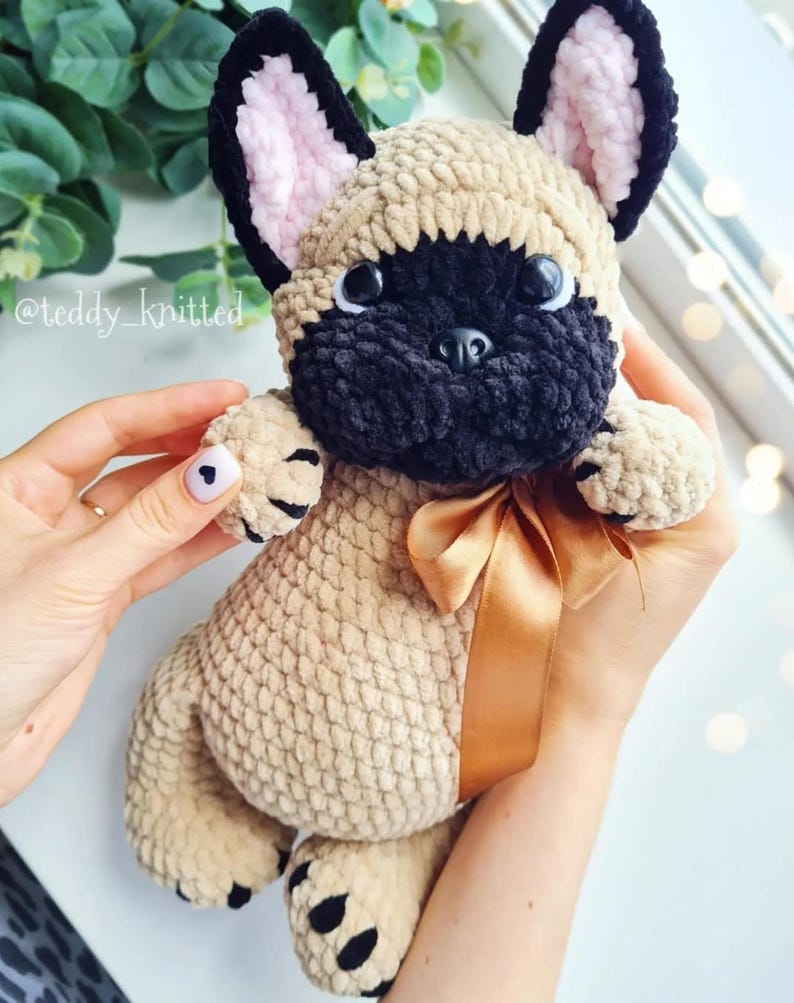May include: A crocheted French bulldog plush toy with a brown and black fur pattern. The toy has a brown ribbon around its neck and black eyes. The toy is being held by a person's hands.