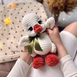 May include: A handmade white crochet duck with a red beak and feet, black eyes, and a green ribbon bow. The duck is a soft, plush toy, perfect for a child's room or as a decorative item. The text "@teddy_knitted" is visible.