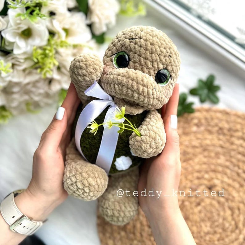 May include: A crocheted plush turtle toy with green eyes, a white ribbon, and a small green flower. The turtle is holding the flower in its right hand. The toy is being held by a person's hands.