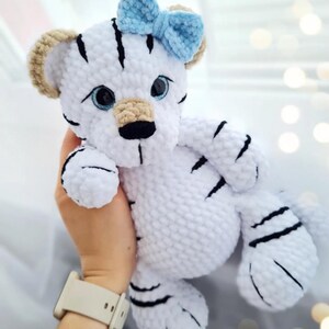May include: A handmade white tiger plush toy with black stripes and a light blue bow. The tiger has tan ears, blue eyes, and a black nose. The plush is soft and cuddly, perfect for a child's toy or nursery decor.