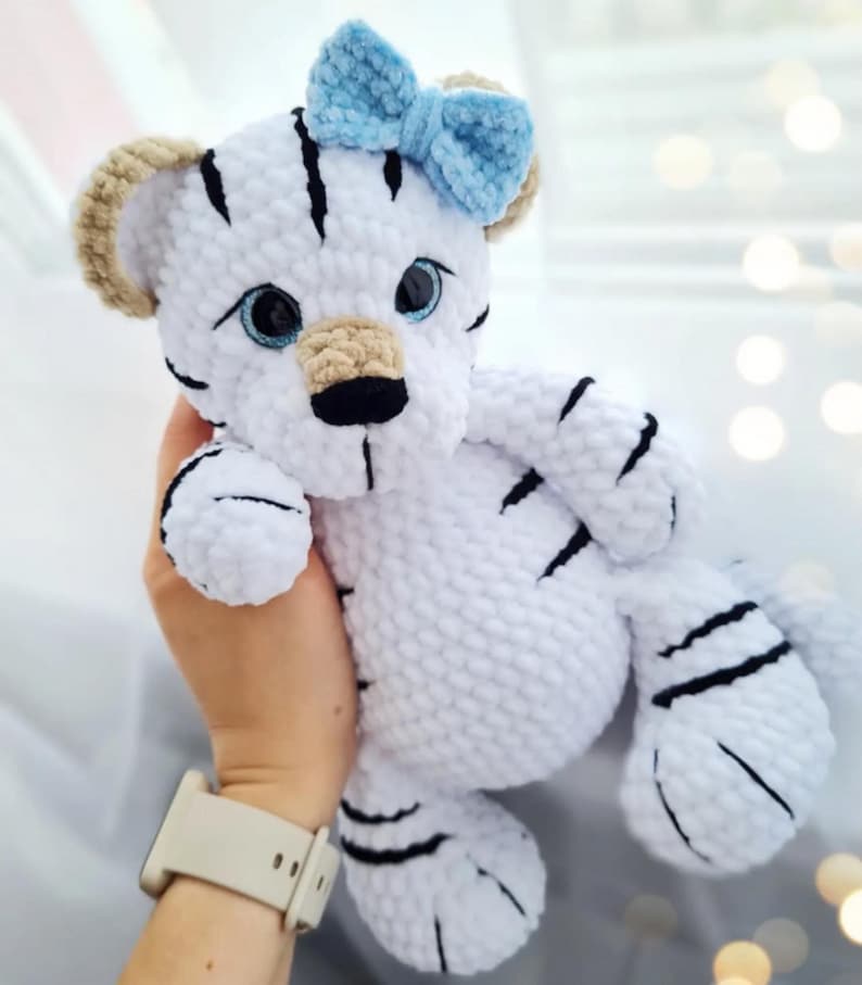 May include: A handmade white tiger plush toy with black stripes and a light blue bow. The tiger has tan ears, blue eyes, and a black nose. The plush is soft and cuddly, perfect for a child's toy or nursery decor.