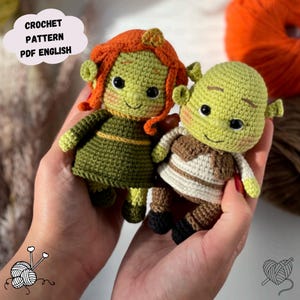 May include: Two handmade crochet dolls, one resembling Shrek and the other Fiona, are held in a person's hands. The Shrek doll is light green with a brown vest, while Fiona has a green dress and orange hair. The text "CROCHET PATTERN PDF ENGLISH" is in a speech bubble.