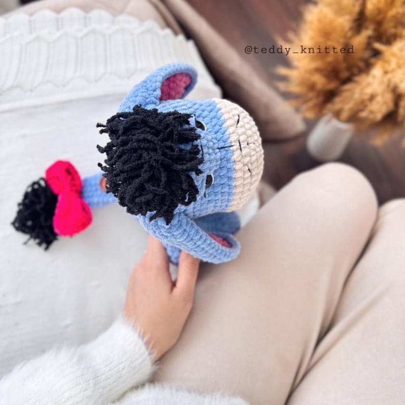 May include: A blue and white crocheted stuffed animal resembling a donkey with a black mane and a pink bow. The stuffed animal is being held by a person wearing a white sweater.