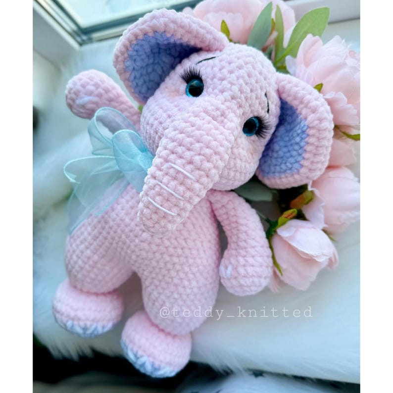 May include: A pink crocheted elephant toy with blue ears and a blue bow. The elephant has embroidered eyelashes and white stitching on its trunk. The toy is sitting on a white fluffy surface.