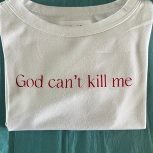 May include: White t-shirt with the text "God can't kill me" printed in red.