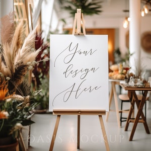 Puede incluir: Un cartel blanco con el texto "Your design here" en un caballete frente a una planta y una mesa con sillas.