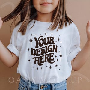 Może przedstawiać: Biały t-shirt z podwiniętymi rękawami z napisem "YOUR DESIGN HERE" w brązowej, retro czcionce, otoczony grafiką gwiazd. Koszulka jest noszona przez osobę w niebieskich dżinsach.