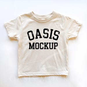 Pode incluir: Uma camiseta de cor creme com texto preto que diz "OASIS MOCKUP" em uma fonte de estilo universitário.