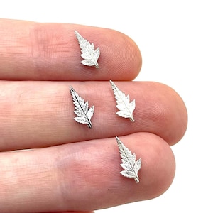 May include: Four small, silver-colored leaf charms are displayed on a fingertip. Each charm has detailed veining and a delicate, textured appearance. The leaves are arranged in a slightly scattered pattern, showcasing their intricate design.
