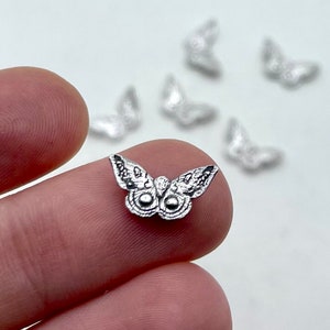 Puede incluir: Primer plano de varios pequeños dijes de mariposa plateados. Uno está enfocado, sostenido entre los dedos, mostrando detalles intrincados. Otros están borrosos en el fondo. Los dijes tienen alas y cuerpo detallados.