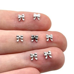 Puede incluir: Primer plano de varios pequeños dijes plateados en forma de lazo. Los dijes están colocados en el dedo de una persona, con un fondo blanco. Los lazos tienen un diseño detallado y probablemente sean componentes de joyería o elementos decorativos.