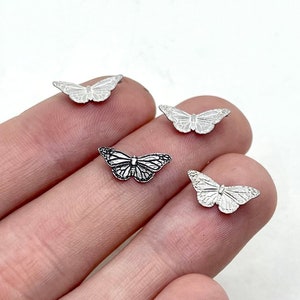 May include: A collection of small, silver-colored butterfly charms. The charms are detailed with wing patterns and are displayed on a light background. One charm has a darker, oxidized finish.