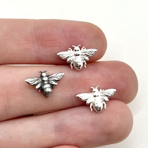 May include: Three small, silver-coloured bee charms with detailed wings and body segments. The bees are displayed on a light background, showcasing their intricate design. These charms are ideal for jewellery making or craft projects.