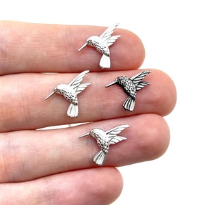 Pode incluir: Cinco pequenos pingentes prateados em forma de beija-flor são exibidos nos dedos de uma pessoa. Os pingentes são detalhados com asas abertas, bicos longos e corpos texturizados. O fundo é branco liso.