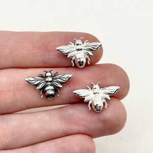 May include: Three small, silver-colored bee charms with detailed wings and body segments. The charms are displayed against a neutral background, showcasing their intricate design. The bees are approximately 0.75 inches in length, perfect for jewelry making or craft projects.