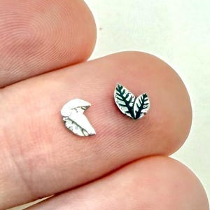 May include: Two small silver-toned stud earrings shaped like leaves. One earring features a smooth, curved leaf design, while the other earring displays a detailed leaf with visible veins and a dark, recessed texture within the leaf pattern.