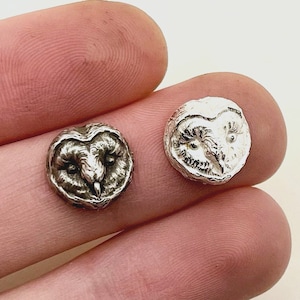 May include: Two small, silver-colored owl-shaped stud earrings. The earrings are round with detailed owl face designs. The owls have large eyes and textured feathers. The earrings are displayed on a light background.