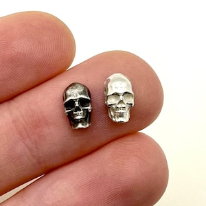 May include: Two small, silver-colored skull charms. One is darker, with a matte finish, and the other is brighter and more reflective. The skulls are detailed with eye sockets, a nose cavity, and teeth. They are likely used for jewelry making or as decorative elements.