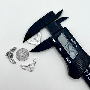 May include: Silver-colored owl charms with outstretched wings, a dime, and a black digital caliper measuring 15.2 mm. The charms are detailed with feathers and facial features. The caliper is set to the "inch/mm" setting.