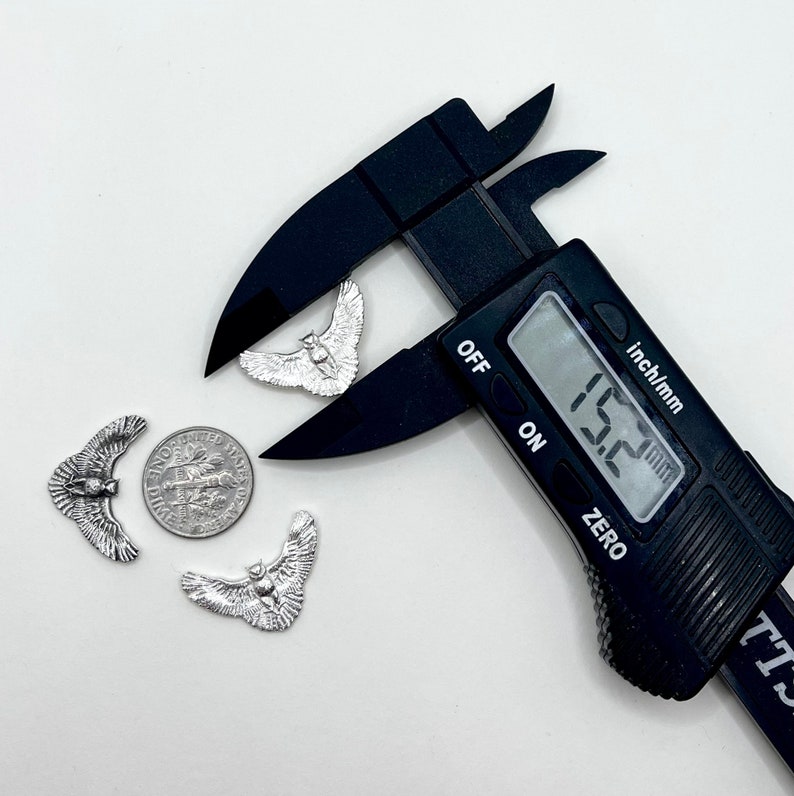 May include: Silver-colored owl charms with outstretched wings, a dime, and a black digital caliper measuring 15.2 mm. The charms are detailed with feathers and facial features. The caliper is set to the "inch/mm" setting.