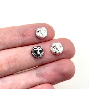 Può includere: Primo piano di tre piccoli ciondoli rotondi color argento. Uno presenta un design dettagliato a testa di ariete, mentre gli altri due hanno un aspetto strutturato e astratto. Questi ciondoli sono probabilmente per la creazione di gioielli o l'artigianato.