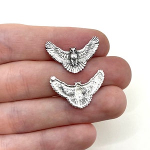 May include: Two silver-colored owl charms with outstretched wings. The charms are detailed with feathers and facial features. The owl charms are small and appear to be made of metal. The charms are held in a hand against a white background.