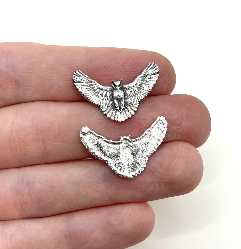 May include: Two silver-colored owl charms with outstretched wings. The charms are detailed with feathers and facial features. The owl charms are small and appear to be made of metal. The charms are held in a hand against a white background.