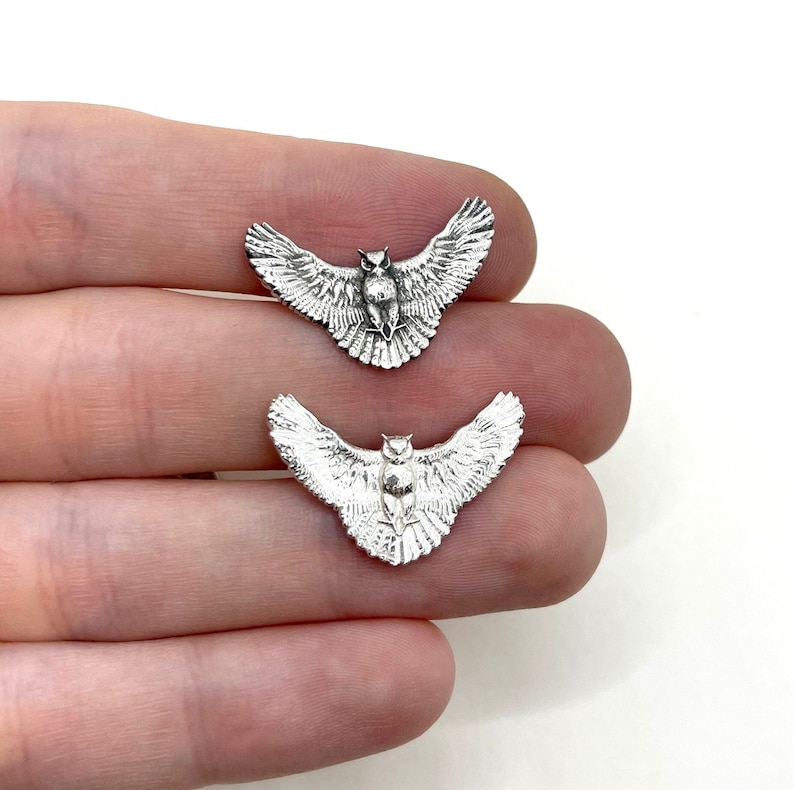 May include: Two silver-colored owl charms with outstretched wings. The charms are detailed with feathers and facial features. The owls are small, and the wings are spread wide. The charms are likely for jewelry making or crafting.