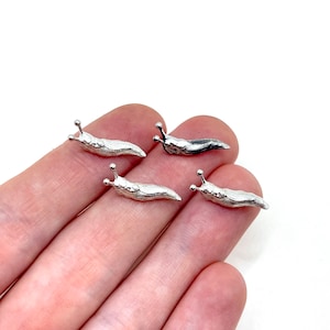 May include: Four small, silver-colored slug figurines. Each slug has a textured body and two antennae with small ball-shaped tips. The slugs are arranged on a light background.