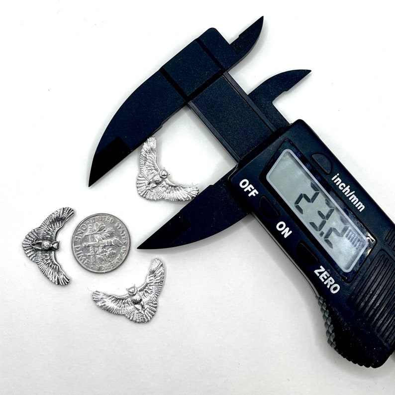 May include: Silver-colored metal owl charms with outstretched wings, displayed with a dime and a digital caliper. The caliper reads 23.2 inches. The charms are detailed with feathers and a central owl body.