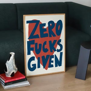 May include: A framed art print with the text "ZERO FUCKS GIVEN" in blue letters over a red heart. The print is displayed on a wooden floor next to a dark green sofa. A small blue lamp sits on a black side table.