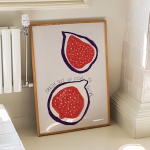 Könnte beinhalten: Ein gerahmter Kunstdruck mit zwei Feigenhälften mit rotem Fruchtfleisch und lila Umrissen auf weißem Hintergrund. Der Text "FRESH OUT OF FIGS TO GIVE" ist um die untere Feige geschrieben. Das Kunstwerk befindet sich in einem Raum mit Fenster, Heizkörper und Buch.