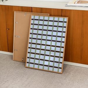 May include: A framed artwork with a geometric pattern in shades of blue, green, and purple. The artwork is leaning against a wall next to an empty frame. The artwork has a light wood frame.